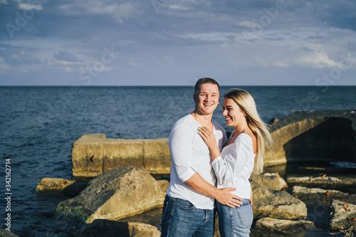 Long shot. Attractive couple in bright clothes standing on stones in the sea against a blue cloudy sky. Lit by the soft evening sun. Looking forward, hug, kissing and enjoying beautiful evening