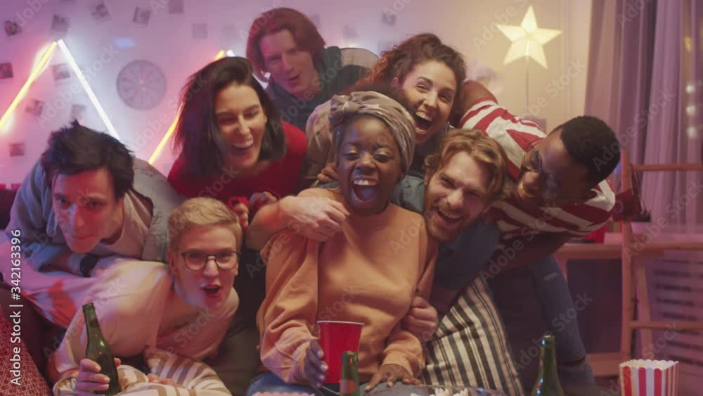Group of joyous young friends hugging cheerful African American woman, smiling and yelling while posing together for camera at home party Group of joyous young friends hugging cheerful African American woman, smiling and yelling while posing together for camera at home party