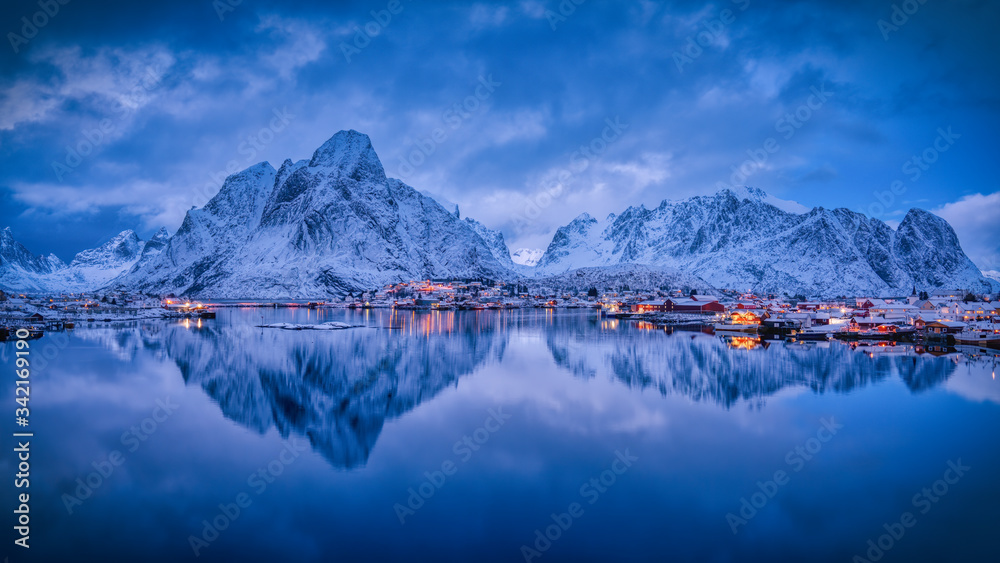 The evening lights of the village of Reine, with snow covered mountain range in the back, and its reflection in the calm water of the fjord.