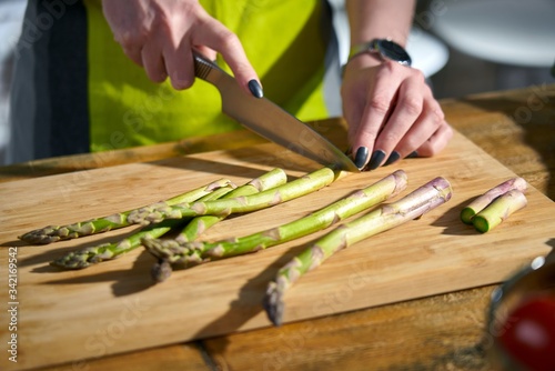 Close-up image of woman cooking in the kitchen, cutting fresh green asparagus on wooden cutting board.