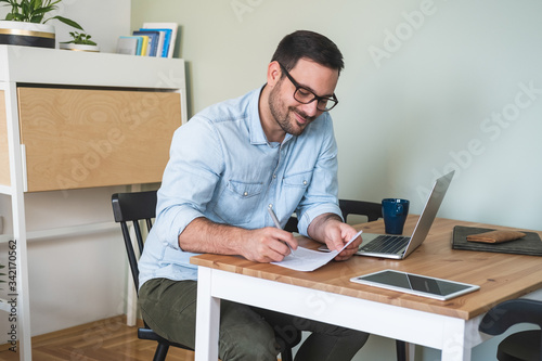 Smiling man sitting at a table with laptop and tablet, writing on paper stock photo