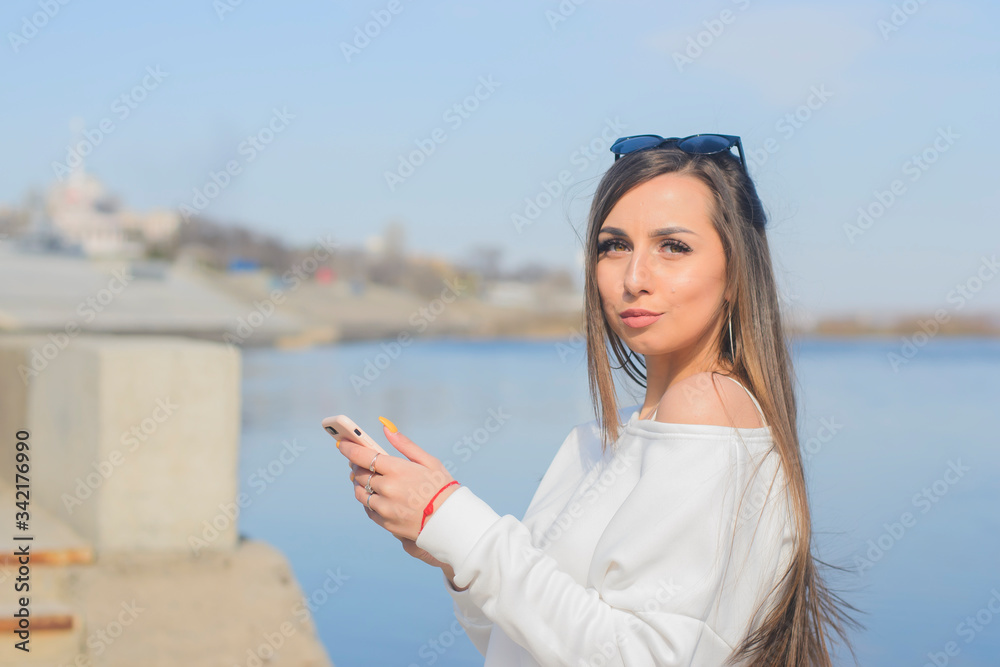 a young girl in a white sweater and sun-protection glasses on her head, with a phone in her hand, walk along the city promenade, and communicate with her partner on the phone