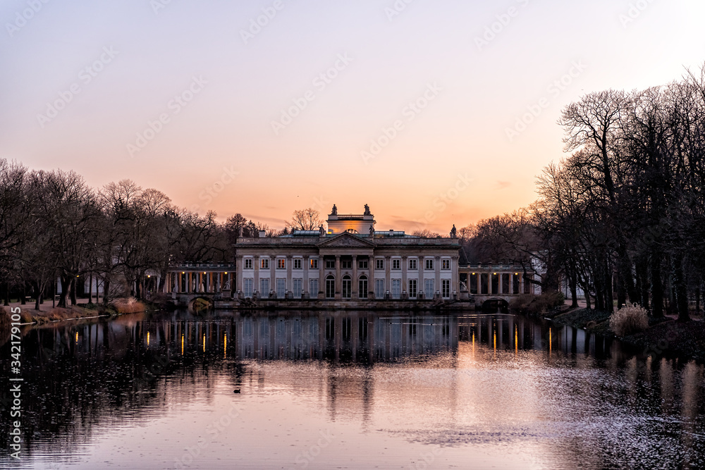 Warszawa Lazienki or Royal Baths Park garden with palace building in center by water lake at sunset in Warsaw, Poland at sunset dusk or twilight