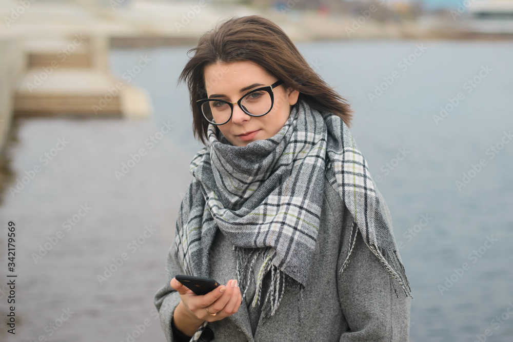 a young girl in glasses, of European descent, in a plaid scarf and a gray coat dials a smartphone number. A girl walks along the embankment of a river or sea
