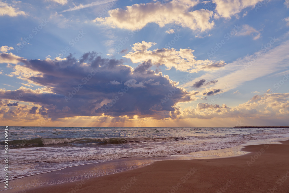 The Sun behind the huge cloud over waving sea and reflections of sky on wet sand on tropical beach.