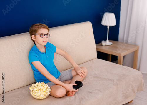 a 9-year-old boy in a blue T-shirt and big glasses sitting on the couch and playing at home with a gamepad. Blue background and free space for text