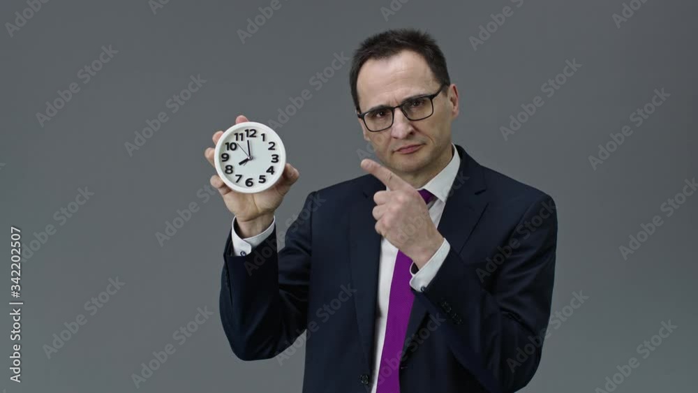 Handsome middle aged businessman in formal clothes, tie and glasses pointing at clock in his hand with time 8 am, on isolated grey background, deadline, time management, working overtime concept Handsome middle aged businessman in formal clothes, tie and glasses pointing at clock in his hand with time 8 am, on isolated grey background, deadline, time management, working overtime concept
