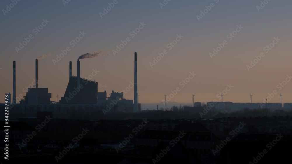 Silhouette of a powerplant in Copenhagen turned into a skislope, with a tall smoking chimney, in sunrise in calm weather with windturbines and the sea in the background. Silhouette of a powerplant in Copenhagen turned into a skislope, with a tall smoking chimney, in sunrise in calm weather with windturbines and the sea in the background.