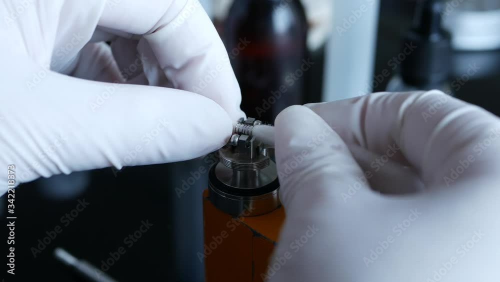 Close up shot of hands wearing white latex gloves inserting wool wick into eCigarette atomizer, rebuilding the coil at home. Close up shot of hands wearing white latex gloves inserting wool wick into eCigarette atomizer, rebuilding the coil at home.