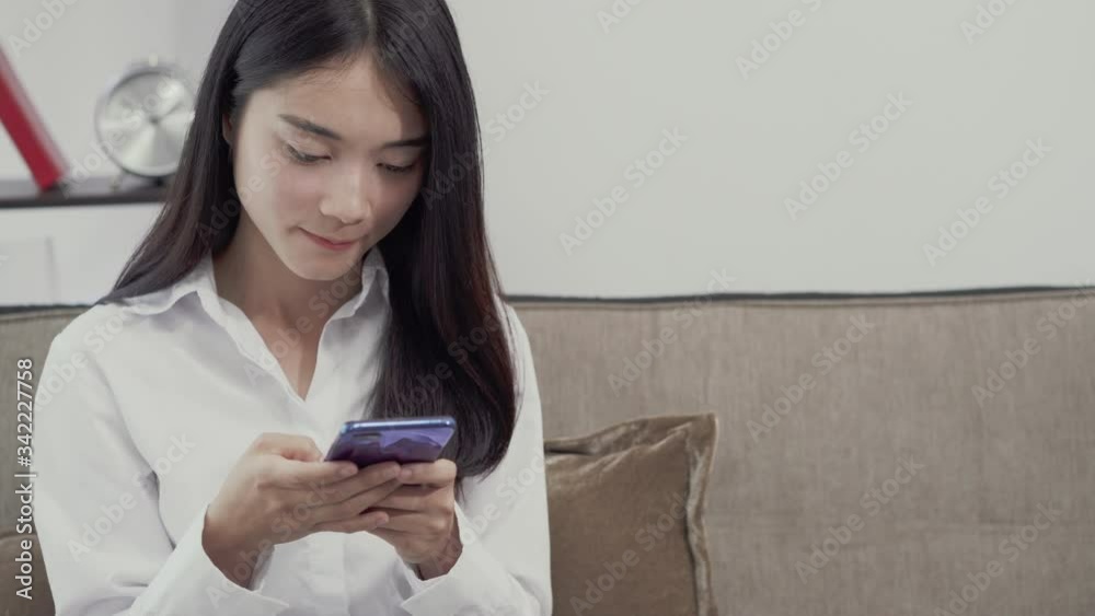 young Asian woman holding smart phone looking at cellphone screen laughing enjoying using mobile apps for shopping or having fun playing games chatting in social media sit at home. work from home young Asian woman holding smart phone looking at cellphone screen laughing enjoying using mobile apps for shopping or having fun playing games chatting in social media sit at home. work from home