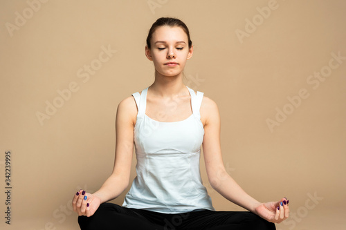 portrait of beautiful young woman working out against beige wall, resting after doing yoga exercises, sitting in ardha Padmasana, Lotus pose, relaxing
