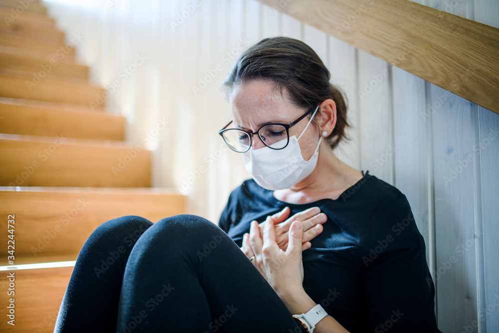 Woman with face mask and chest pain sitting indoors at home, Corona virus and quarantine concept.