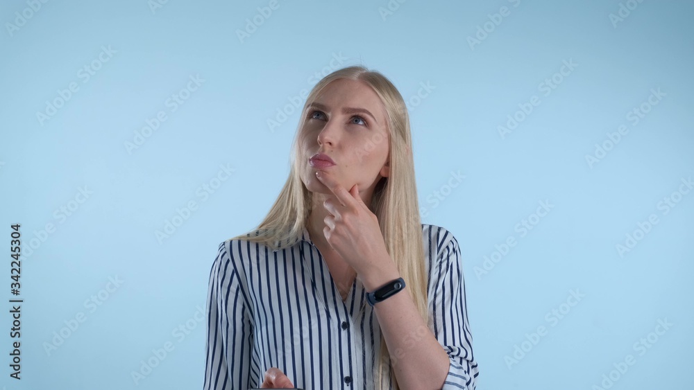 Blonde young woman holding her chin with hand while thinking about something on blue background. She looking at smartphone and making plans.