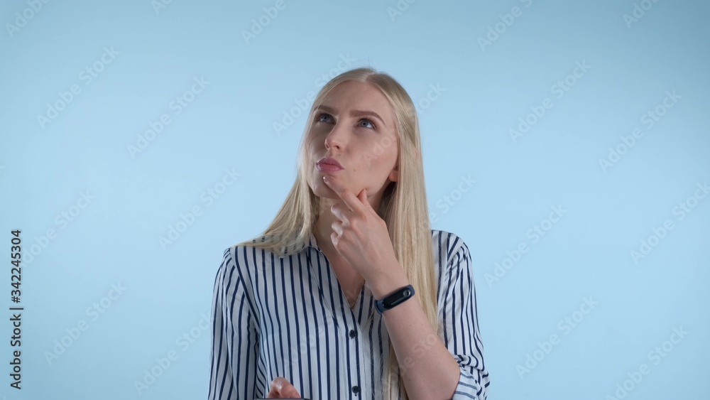Blonde young woman holding her chin with hand while thinking about something on blue background. She looking at smartphone and making plans. Blonde young woman holding her chin with hand while thinking about something on blue background. She looking at smartphone and making plans.