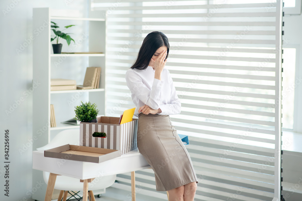 Upset young brunette female sitting on her desk, closing her face with one hand Upset young brunette female sitting on her desk, closing her face with one hand