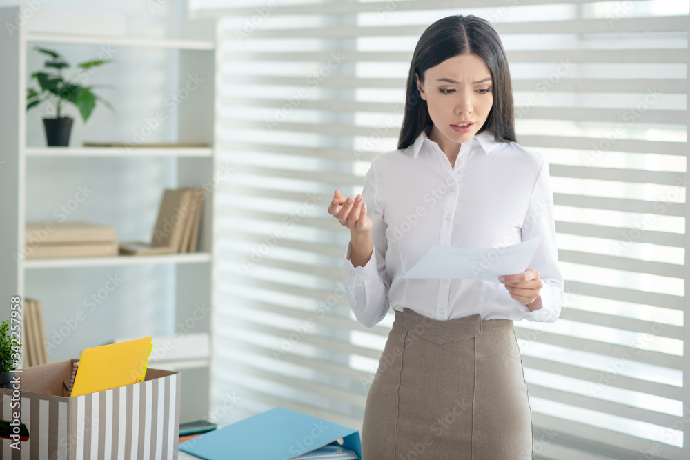 Upset young brunette female holding piece of paper, reading it Upset young brunette female holding piece of paper, reading it