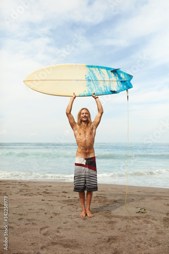 Surfing. Handsome Young Surfer Holding White Surfboard Above Head. Smiling Man Standing On Sandy Beach Portrait. Beautiful Ocean And Blue Sky With Soft Clouds Background.