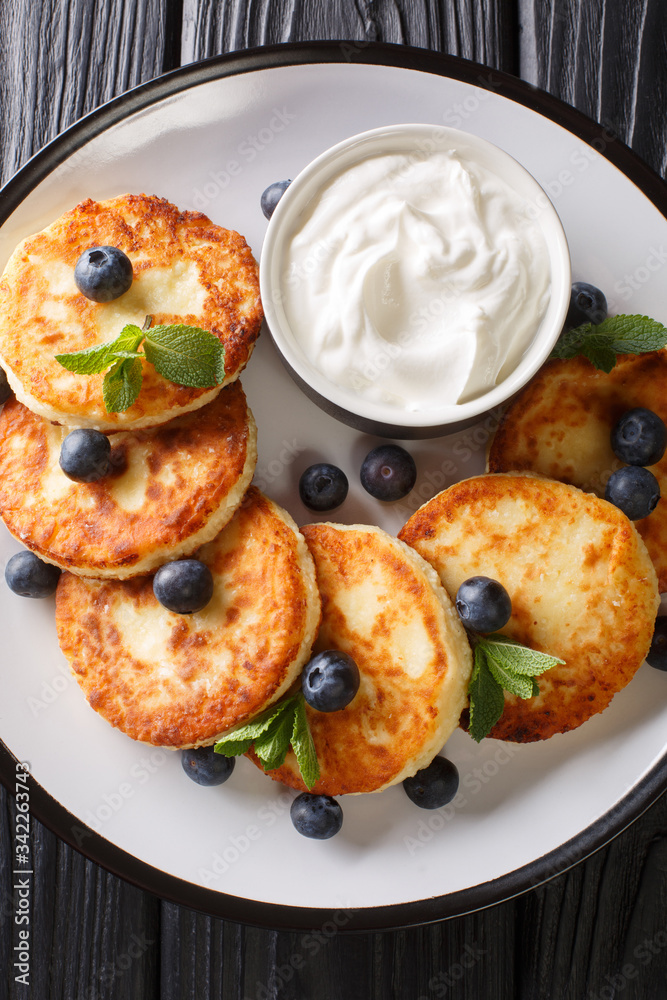 Tasty syrniki with blueberries, mint and sour cream close-up in a plate. Vertical top view Tasty syrniki with blueberries, mint and sour cream close-up in a plate. Vertical top view