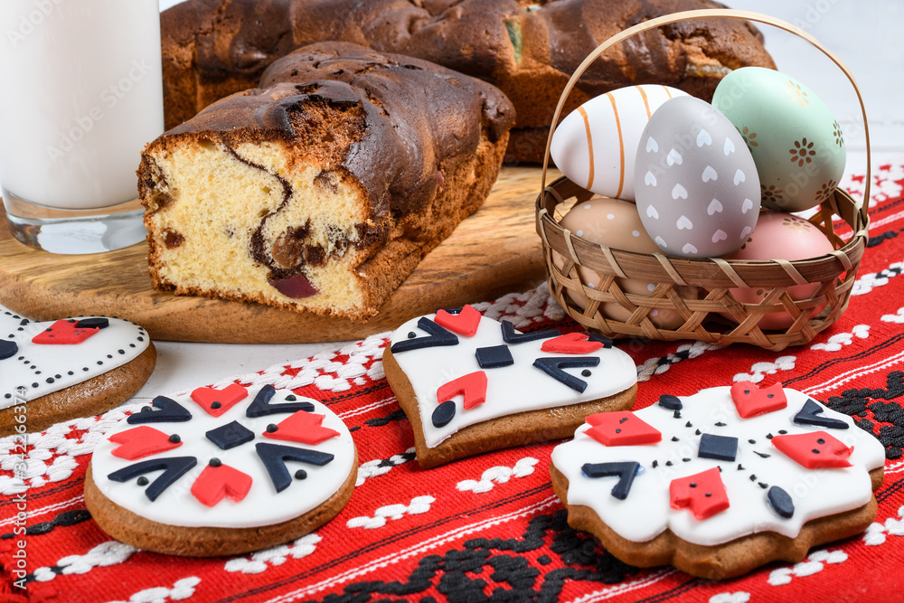 Slices of Romanian sponge cake or chec, cozonac with red traditional towel with manual stitch on wooden board, Easter painted eggs and home made ginger bread hearts, Authentic Easter Brunch concept. 