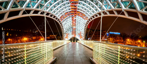 Fototapeta Naklejka Na Ścianę i Meble -  Panoramic image of architecture and pathway of iliuminated Peace bridge in Tblisi at night time. Georgia.25.03.2020.
