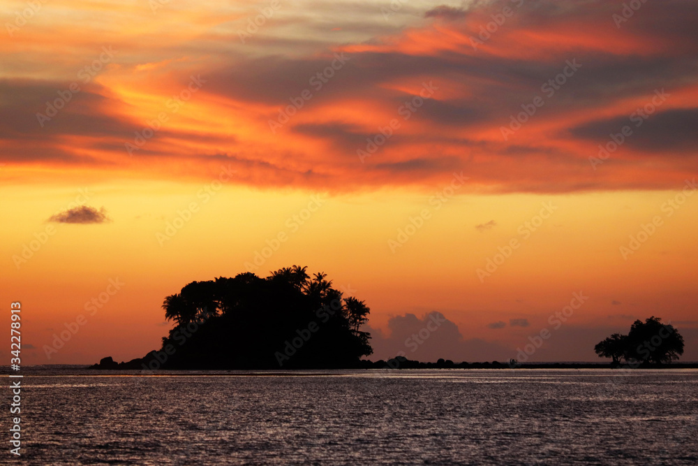 Sunset on a beach, silhouette of tropical island with palm trees in dark sea. Dramatic orange sky with clouds, romantic vacation concept Sunset on a beach, silhouette of tropical island with palm trees in dark sea. Dramatic orange sky with clouds, romantic vacation concept
