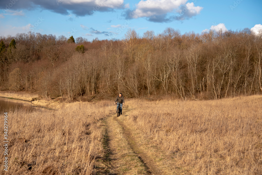 A man rides a Bicycle on dry grass against a background of bare trees.