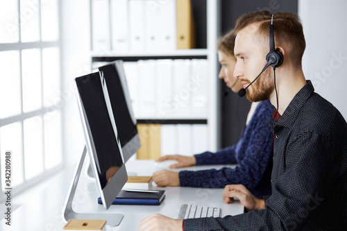 Red-bearded businessman talking by headset near his female colleague while sitting in modern office. Diverse people group in call center. Telemarketing and customer service