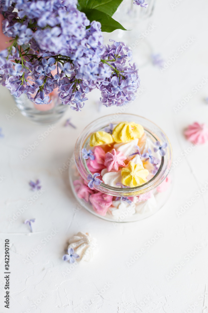 Some meringue kisses in a glass jar on the wait background decorated with bouquet of fresh lilac flowers. Spring season. Some meringue kisses in a glass jar on the wait background decorated with bouquet of fresh lilac flowers. Spring season.