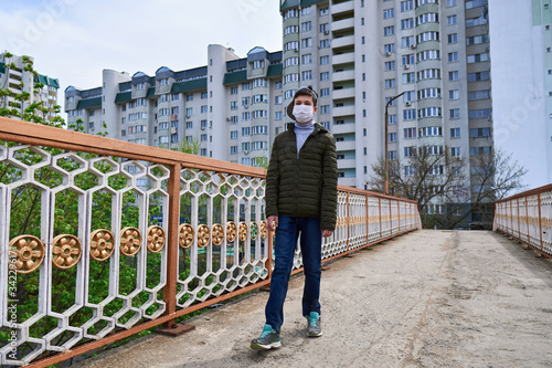 teen boy walks down the street during the day, a pedestrian walkway and high-rise buildings with apartments, a residential area, a medical mask on his face protects against viruses and dust © soleg