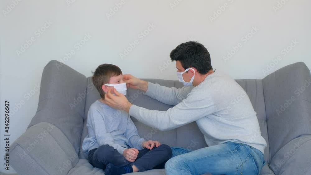 Dad is teaching son putting on mask on face sitting on couch at home, coronavirus pandemic quarantine. Worldwide global COVID-19 epidemic. Fighting with 2019-ncov infection. Family protection. Dad is teaching son putting on mask on face sitting on couch at home, coronavirus pandemic quarantine. Worldwide global COVID-19 epidemic. Fighting with 2019-ncov infection. Family protection.