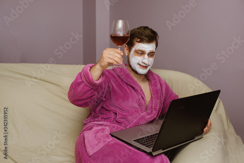Photo of happy smiled funny man wearing pink robe with facial masks while having video call on laptop at home home isolation quarantine