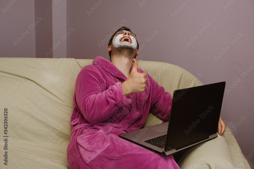 Happy laughing man with facial mask wearing pink robe sitting on the sofa and speaking with his friends on laptop at home. Man watching funny movie on laptop during the home isolation quarantine Happy laughing man with facial mask wearing pink robe sitting on the sofa and speaking with his friends on laptop at home. Man watching funny movie on laptop during the home isolation quarantine