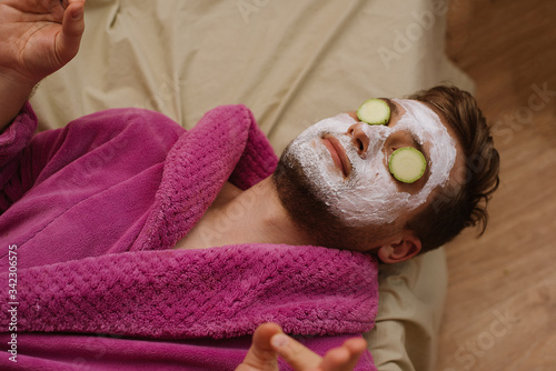 Close up photo of relax meditate man wearing pink robe with facial mask and cucumber on his face laying on the sofa at home isolation quarantine. spa at home concept