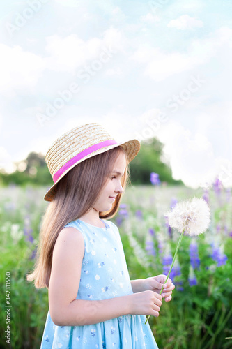 Beautiful little girl in a straw hat and dress holds a dandelion in a flowering field. Nature concept. A smiling girl is playing on the summer lawn. Allergy. Childhood. Copy space. Summer joy 