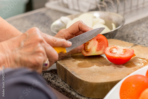 Old woman hands cutting a tomato on wooden board with knife in kitchen.
