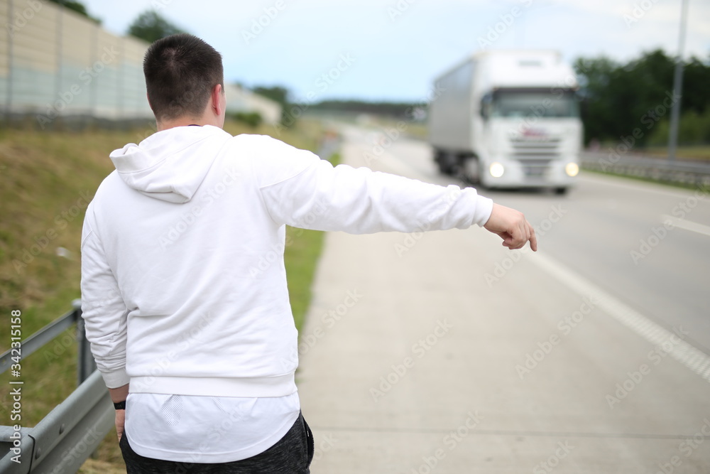 young man in a white pullover walking on the road and trying to stop a car. Young traveler hitchhiking .  Travel by car.