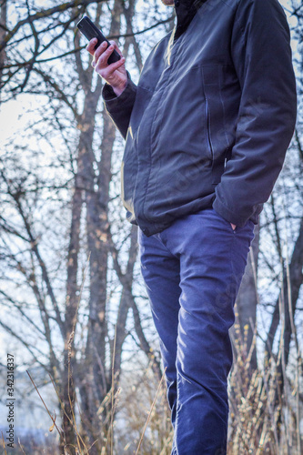 A thin man in a jacket and jeans with a phone in his hands is standing outdoors in a city park in spring