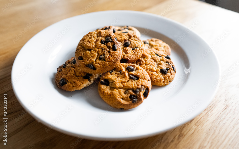 Choc chip cookies in a white plate on a wooden table. Close-up of homemade chocolate chip cookies. Basic drop cookies with chocolate morsels. Traditional handmade chewy cookies with chocolate chunk Choc chip cookies in a white plate on a wooden table. Close-up of homemade chocolate chip cookies. Basic drop cookies with chocolate morsels. Traditional handmade chewy cookies with chocolate chunk