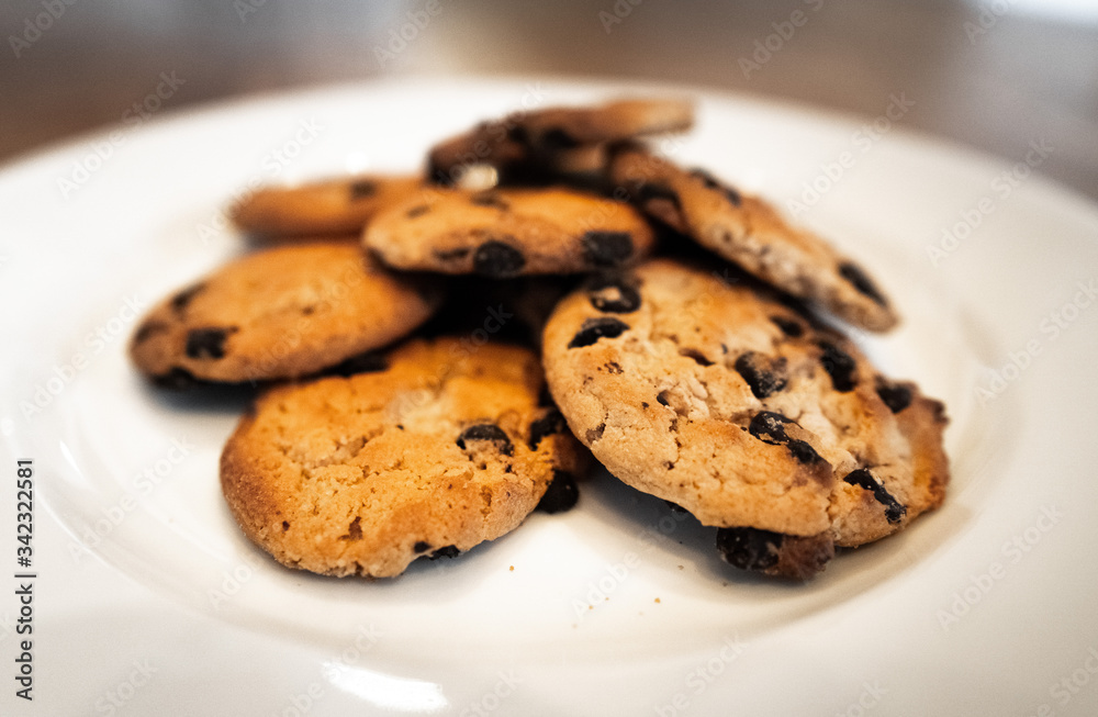 Close-up of choc chip cookies in a white plate on a wooden table. Homemade chocolate chip cookies. Basic drop cookies with chocolate morsels. Traditional handmade chewy cookies with chocolate chunk Close-up of choc chip cookies in a white plate on a wooden table. Homemade chocolate chip cookies. Basic drop cookies with chocolate morsels. Traditional handmade chewy cookies with chocolate chunk