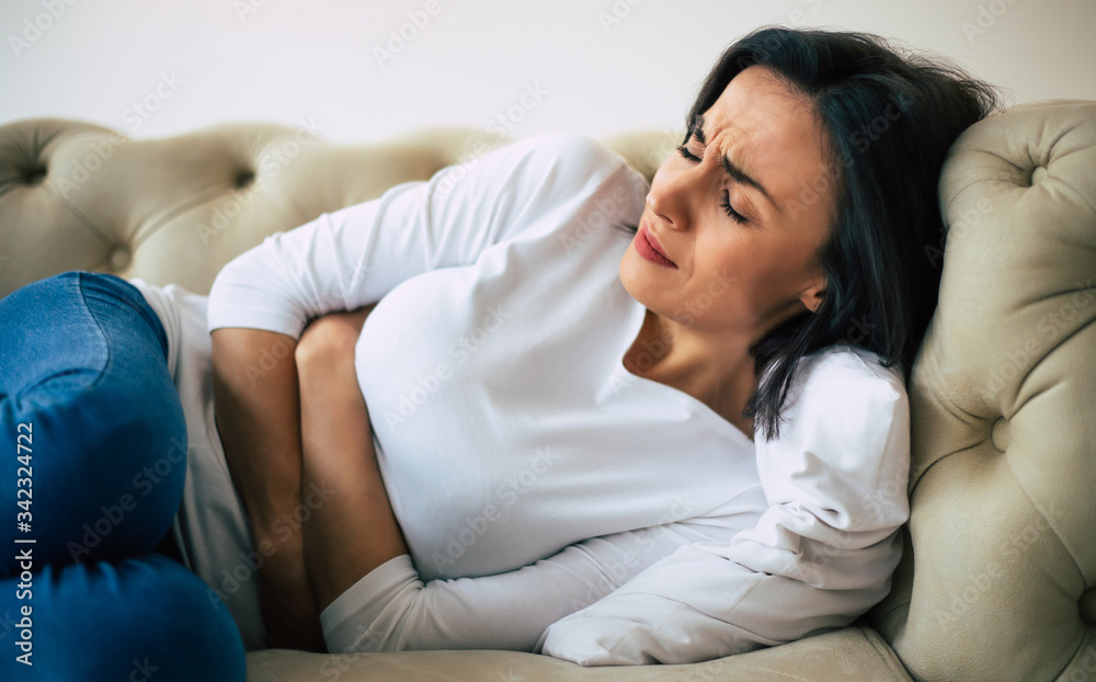 Dysmenorrhea. Close-up photo of a woman, who is lying on her sofa and holding her stomach with suffering facial expression. Dysmenorrhea. Close-up photo of a woman, who is lying on her sofa and holding her stomach with suffering facial expression.