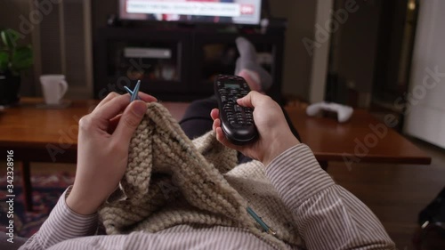 Woman changes the channel on the TV with a remote while crocheting on the couch