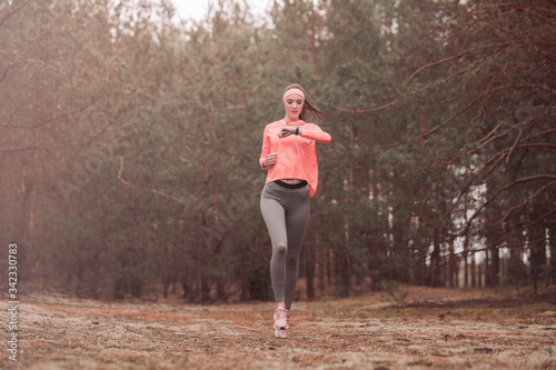 Woman running in the woods and checking smartwatch. Young girl jogging in a forest. Healthy lifestyle fitness sporty woman running early in the morning. Beautiful young woman runner in a pine forest.