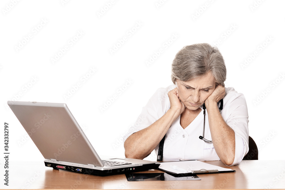 Serious elderly woman doctor sitting at table with computer isolated Serious elderly woman doctor sitting at table with computer isolated