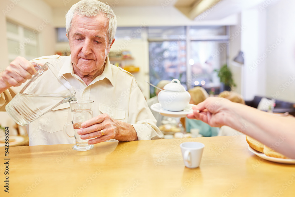 Senior gießt Wasser aus einer Karaffe in ein Glas im Aufenthaltsraum im Seniorenheim Senior gießt Wasser aus einer Karaffe in ein Glas im Aufenthaltsraum im Seniorenheim