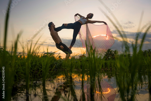 outdoors sunset acroyoga workout - young happy and fit couple practicing acro yoga drill at beautiful rice field enjoying nature doing acrobatic pose with woman in wings