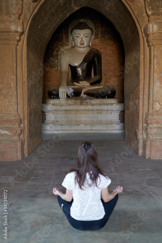 A female is meditating in lotus position, sitting on the floor in front of a Buddha statue in an ancient temple in Bagan in Myanmar.