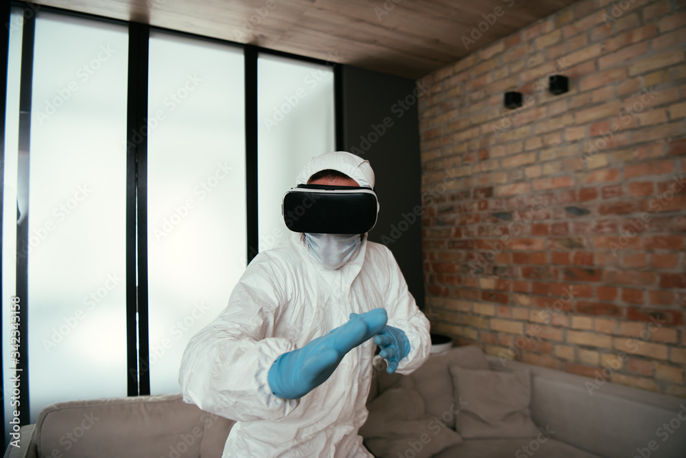 man in hazmat suit, medical mask, latex gloves and virtual reality headset gesturing in living room