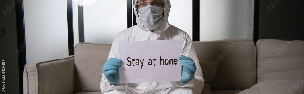 panoramic shot of man in personal protective equipment and latex gloves holding paper with stay at home lettering