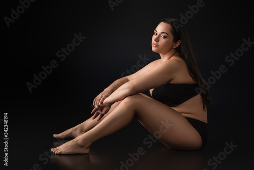 Side view of beautiful plus size girl in underwear sitting on black background
