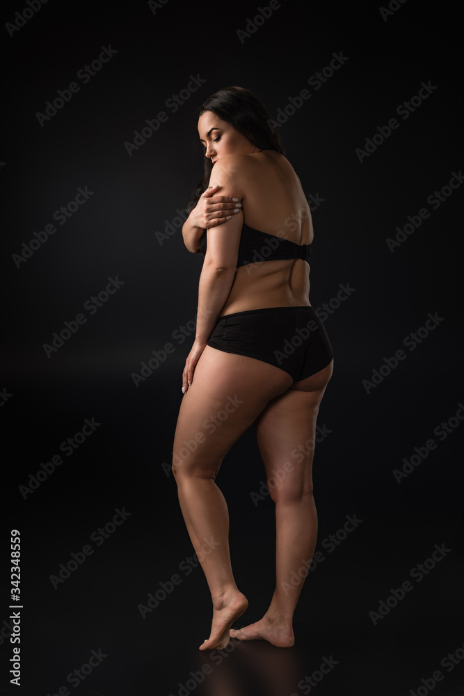 Fototapeta premium Full length view of plus size girl hiding body with shame on black background